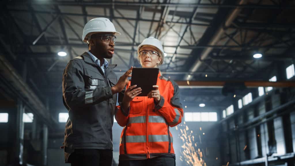Two Engineers Stand in a Manufacturing Factory, while using a Digital Tablet Computer and Having a Discussion.